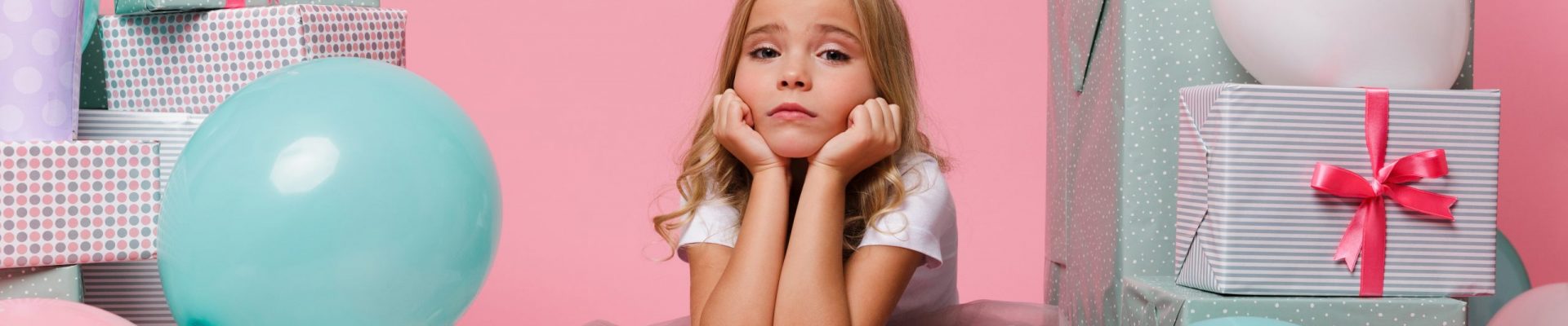 Portrait of a sad pretty little girl in a birthday hat sitting in a pile of present boxes and looking at camera isolated over pink background