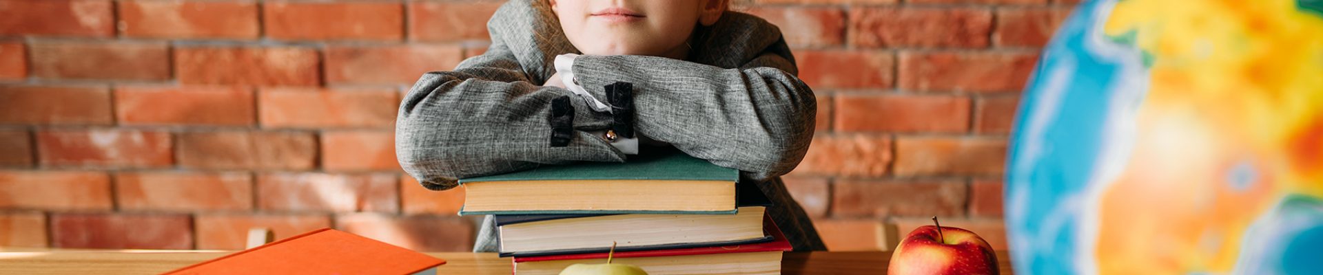 Serious schoolgirl with textbooks poses at the table. Female pupil reading a book at the desk, young girl doing homework