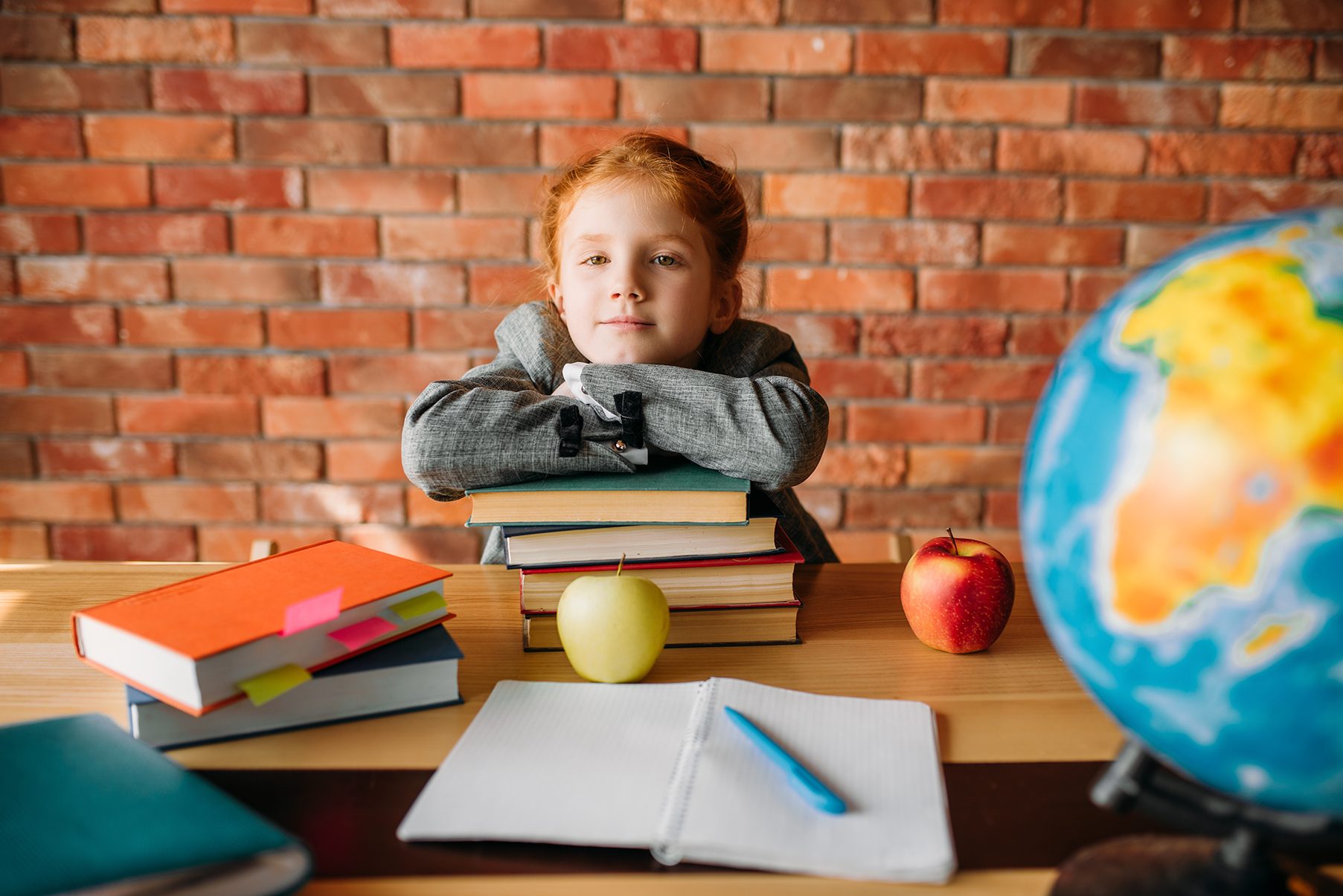 Serious schoolgirl with textbooks poses at the table. Female pupil reading a book at the desk, young girl doing homework