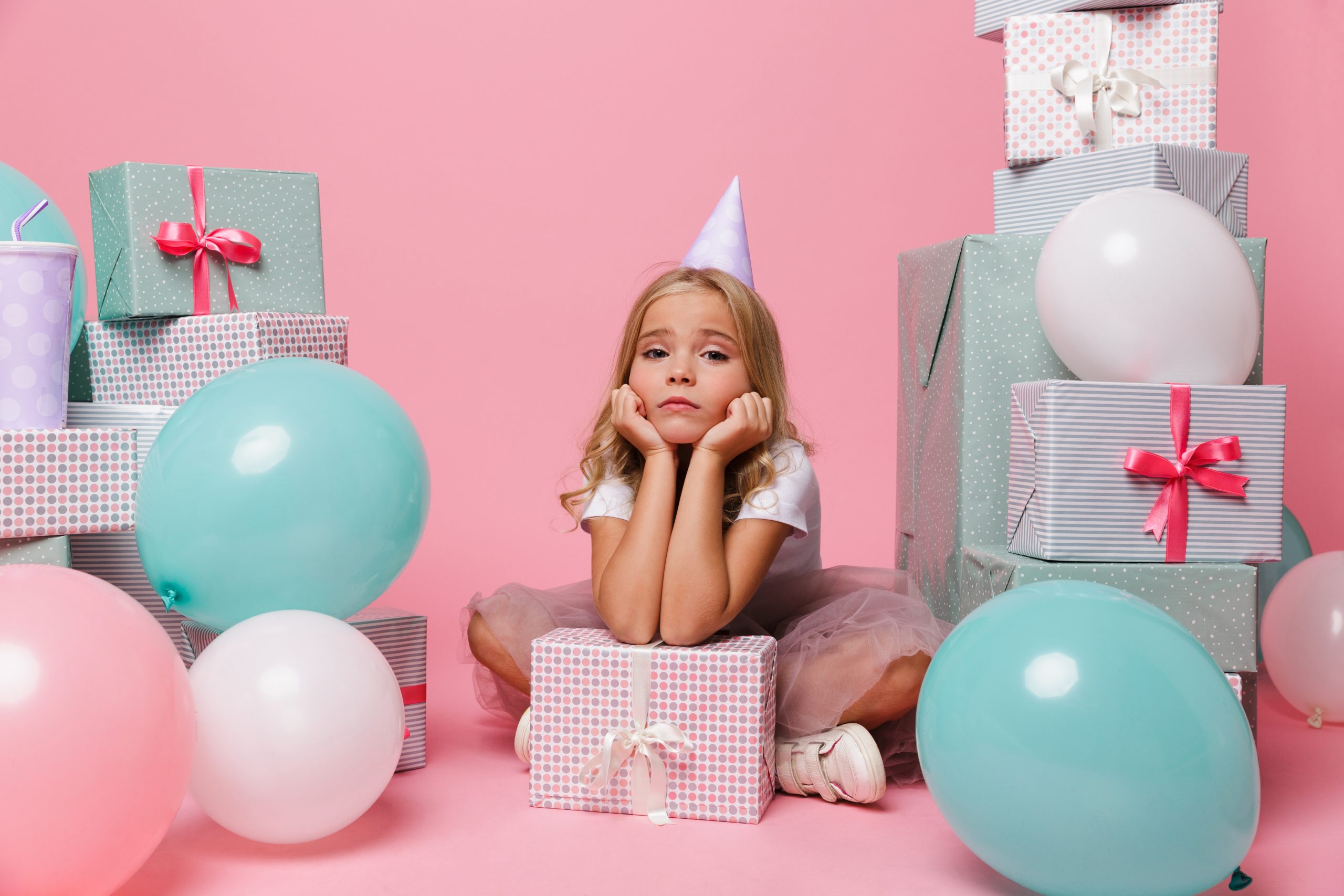 Portrait of a sad pretty little girl in a birthday hat sitting in a pile of present boxes and looking at camera isolated over pink background
